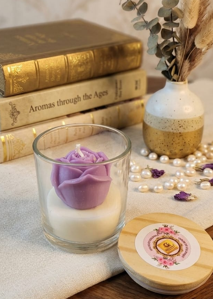 Purple rose Candle with a decorative lid on a wooden surface with books and a vase in the background.