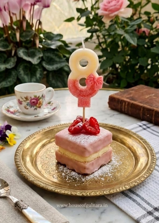 Small pink cake with a number 8 candle on a gold plate, surrounded by flowers and a teacup.