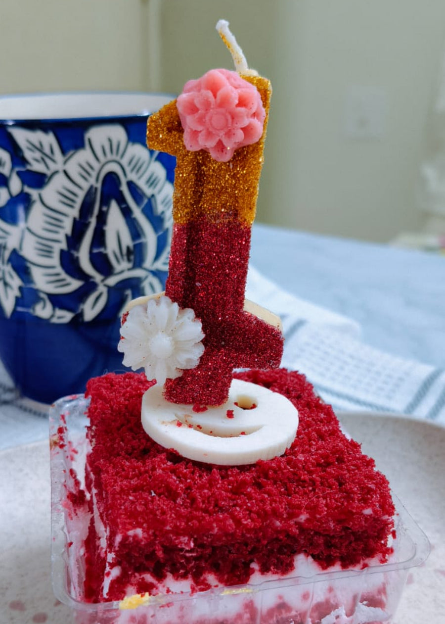 Square slice of red velvet cake with a lit candle on a white plate.