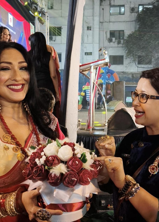 Two women, one holding a bouquet of flowers, in an urban setting with buildings and a colorful structure in the background.