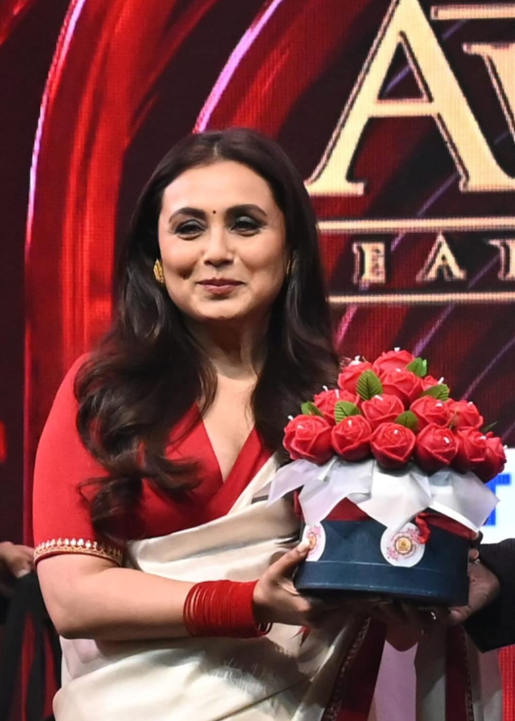 Rani Mukerjee bollywood actress in a red and white saree holding our candle bouquet on a stage with a red and gold background