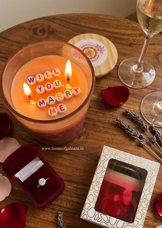 Candle with 'Will You Marry Me' message, ring box, and rose petals on a wooden table.