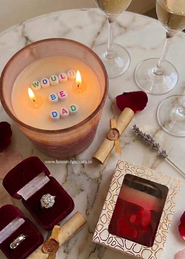 Candle with letter blocks spelling 'WOULD BE DAD', ring box, and wine glasses on a marble surface.