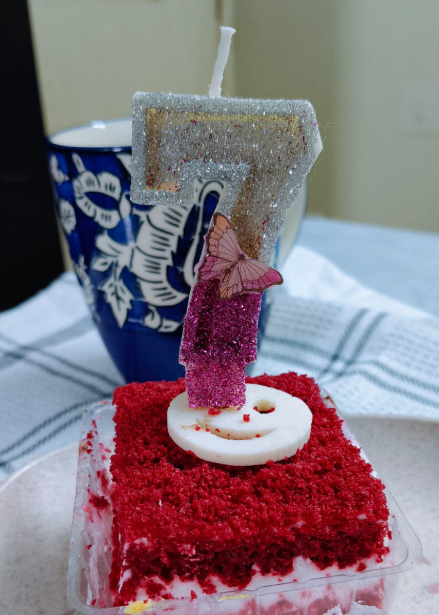 Square piece of red velvet cake with a lit candle on a white plate.