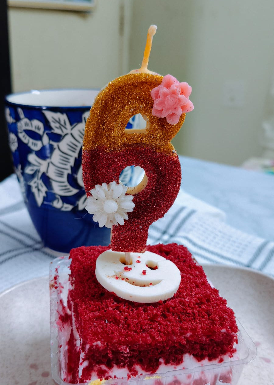 Red velvet cake with a number 6 candle on a white plate, next to a blue mug.