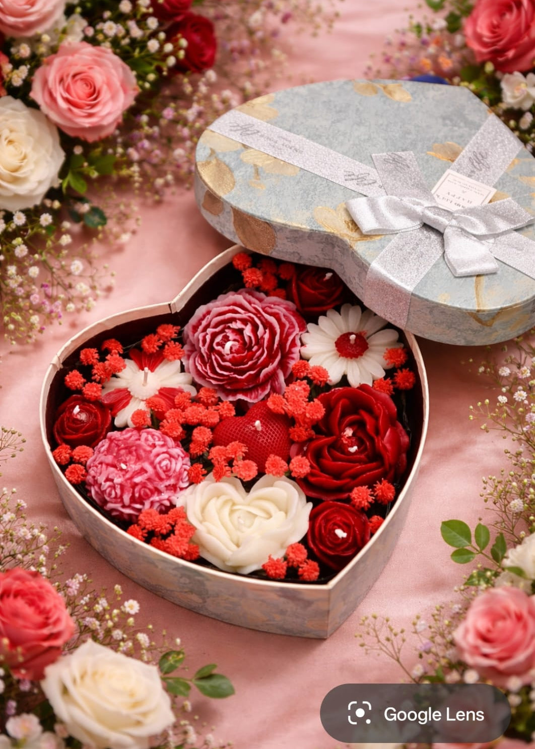 Heart-shaped box filled with red, white, and pink rose and diffrent types of floral scented candles on a pink floral background.