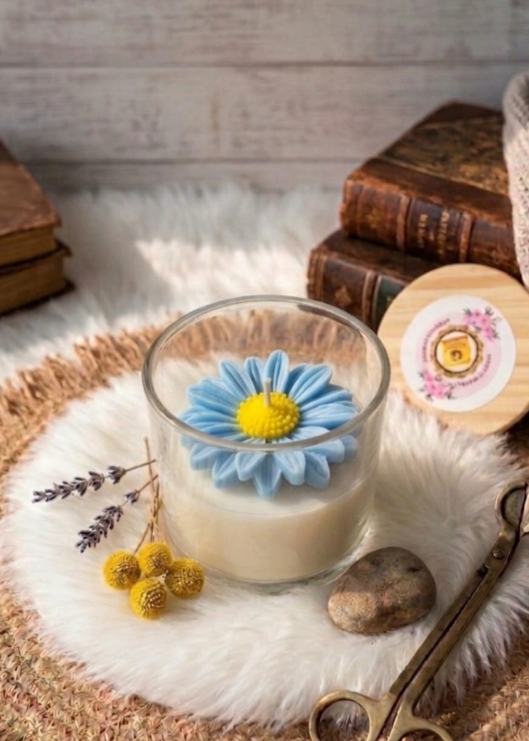 Glass of blue daisy candle with a decorative flower, cookies, and books on a textured surface