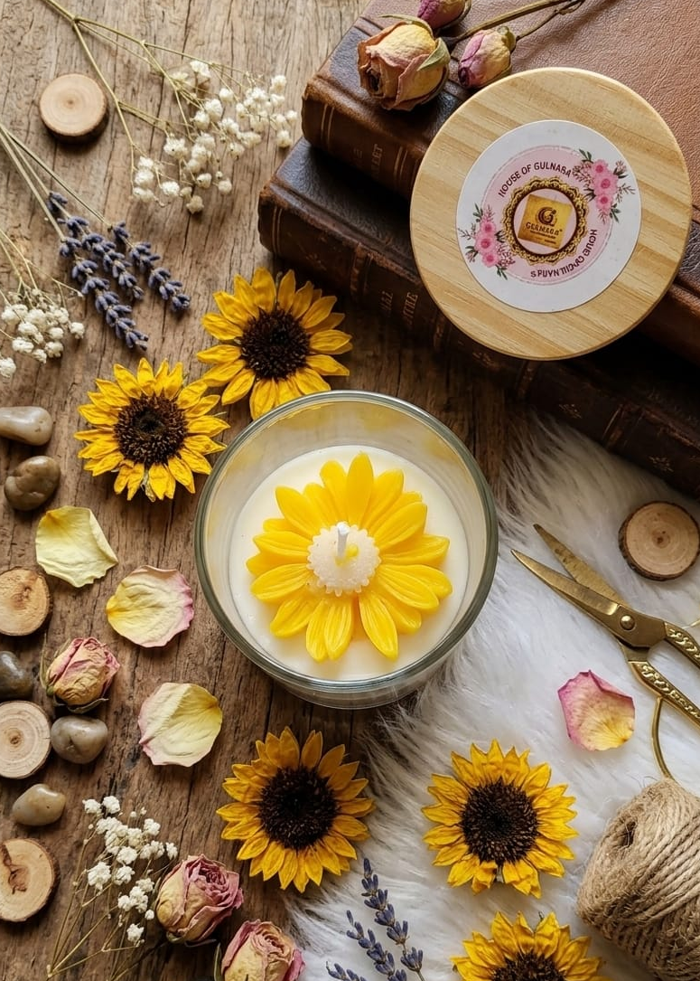 Candle with daisy design surrounded by sunflowers, lavender, and stones on a wooden surface.