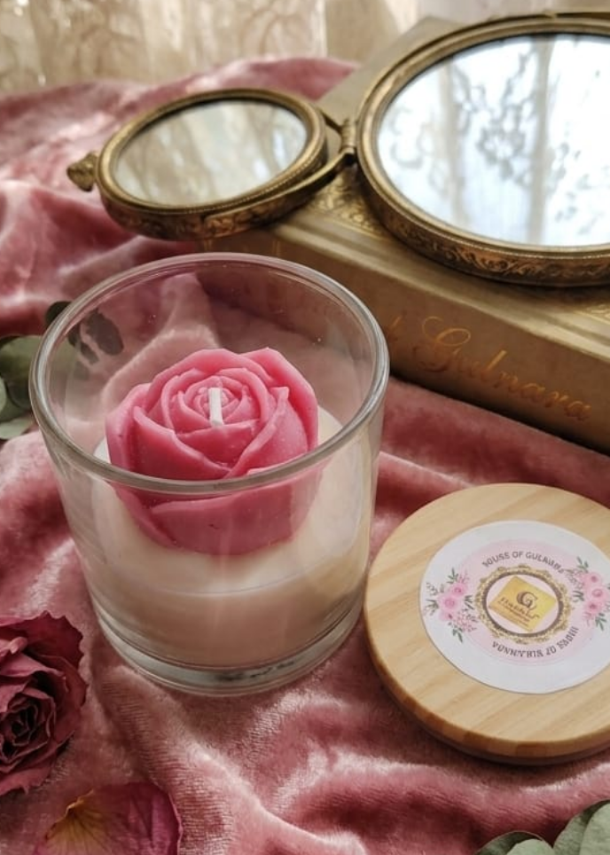 Decorative setup with a pink candle in a glass holder, pink rose, and small round mirror on a pink fabric background.