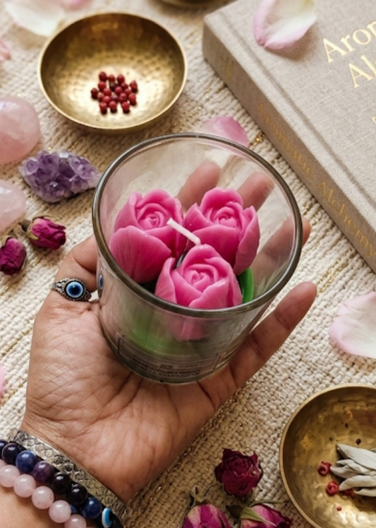 Hand holding a glass with pink floral tulip candles, surrounded by crystals and books on a textured surface.