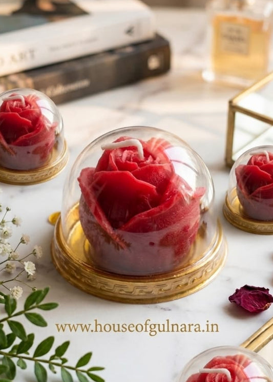 Three red roses in glass domes on a marble surface with books and a plant in the background.