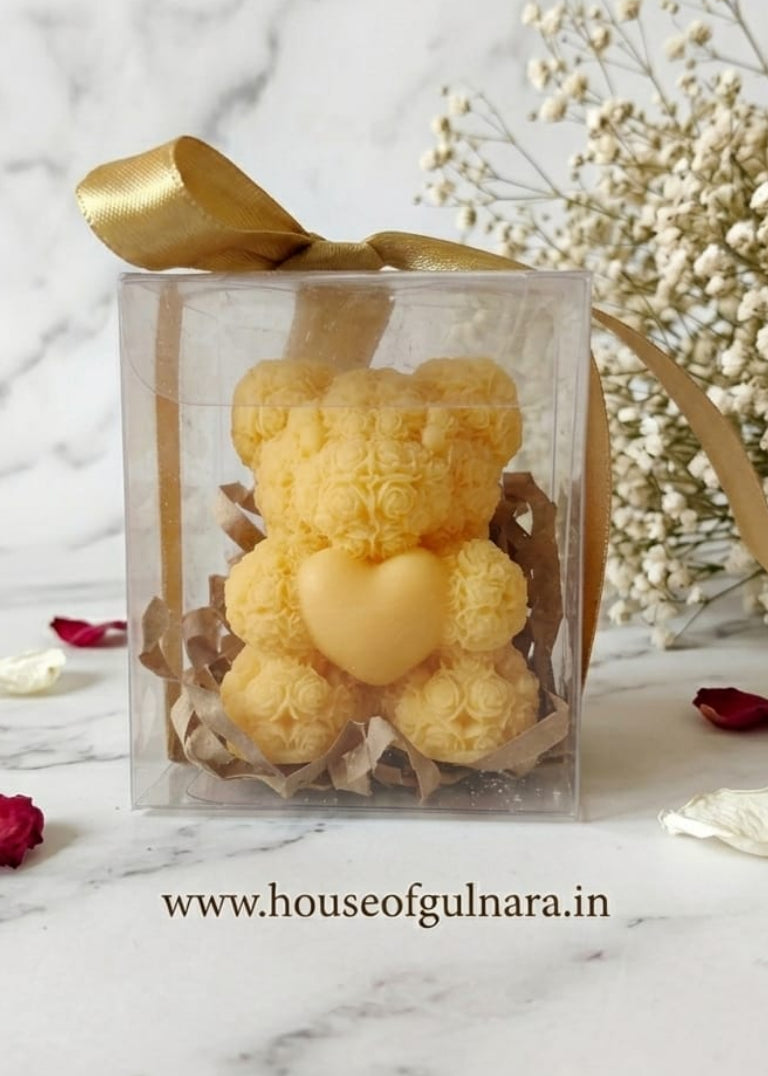 Clear box with a yellow teddy bear candle and gold ribbon on a marble surface with white flowers and red petals.