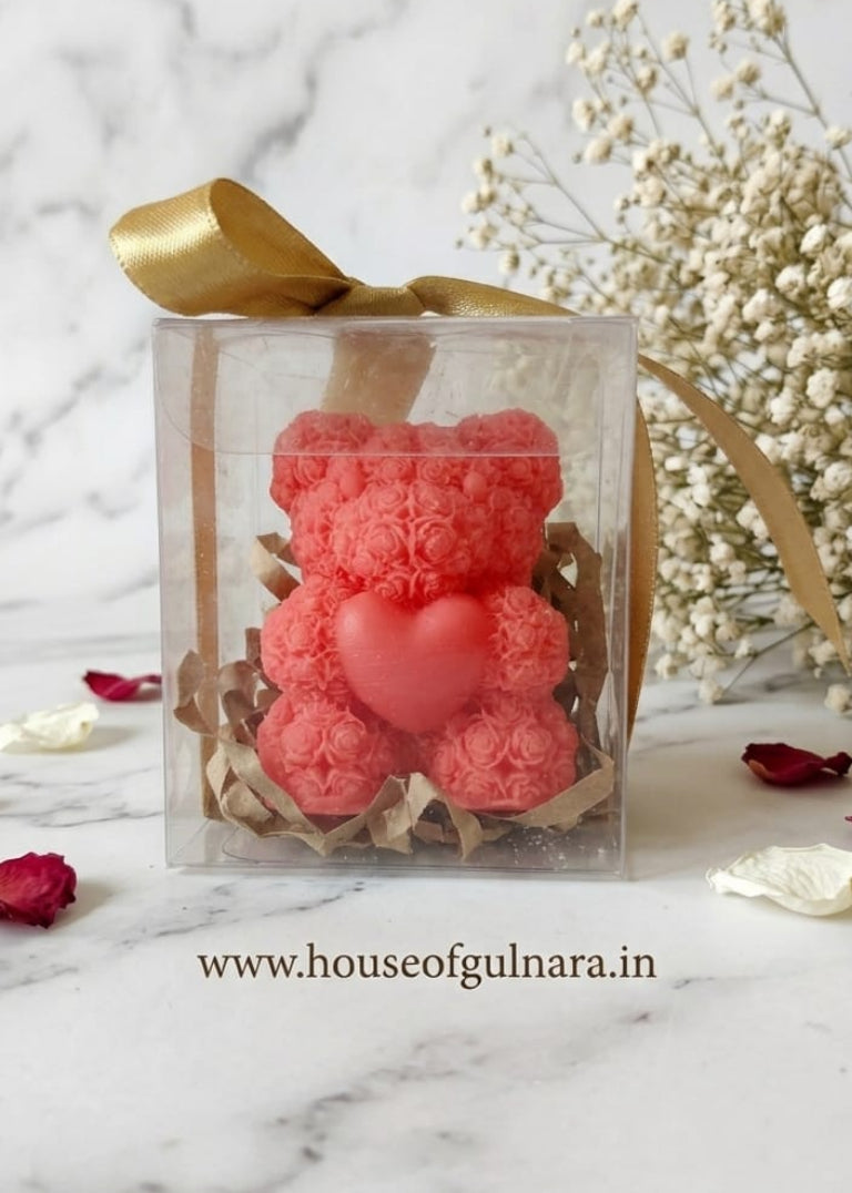 Pink teddy bear-shaped candle in a clear box with gold ribbon on a marble surface.