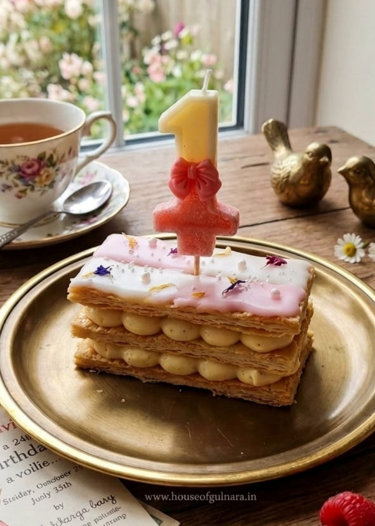 Small cake with a number 1 candle, tea cup, and birthday card on a wooden table with flowers in the background.