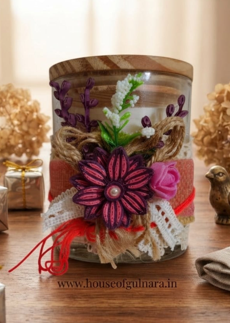 A decorative glass jar candle with a wooden lid, filled with layered dried flowers, burlap, lace, and ribbon, displayed on a wooden table with dried flowers and small wrapped gifts in the background.
