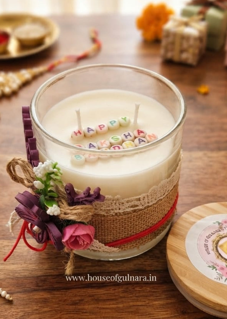 A glass jar candle with white wax sits on a rustic wooden table, decorated with a burlap wrap, red ribbon, and small floral accents. The lid rests beside it, with decorative flowers, jewelry, and wrapped gifts softly blurred in the background.