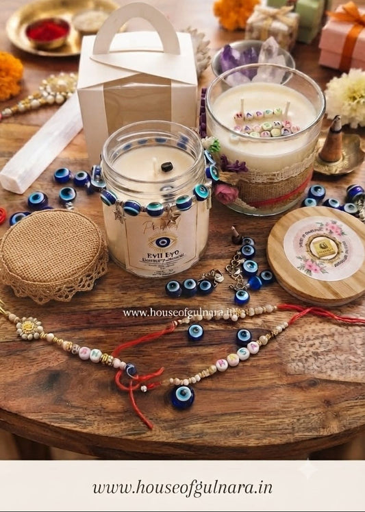 A styled tabletop display featuring a glass jar candle labeled 'Evil Eye' with blue evil eye beads around the rim, a white gift box with a handle, a larger glass candle with floral accents, and additional decorative items including beaded necklaces, a woven lid, and small wrapped gifts on a rustic wooden surface.