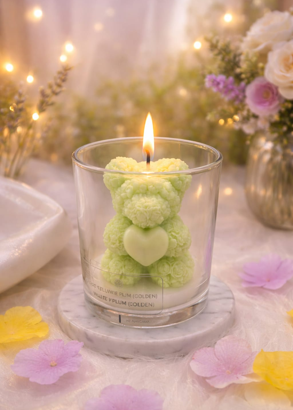 Candle shaped like a green teddy bear with a heart on a white surface with flowers and lights in the background.
