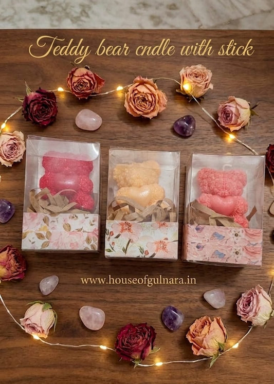 Three teddy bear candles in packaging with decorative flowers and lights on a wooden surface.