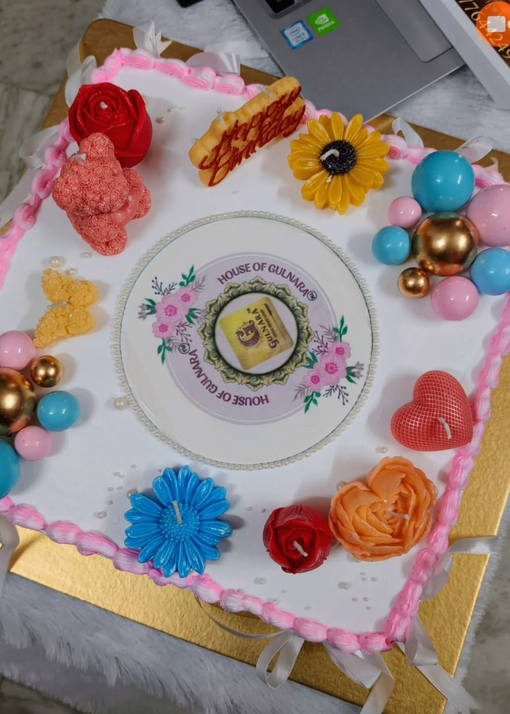 Decorated birthday cake with colorful flowers and balloons on a table with a laptop in the background.