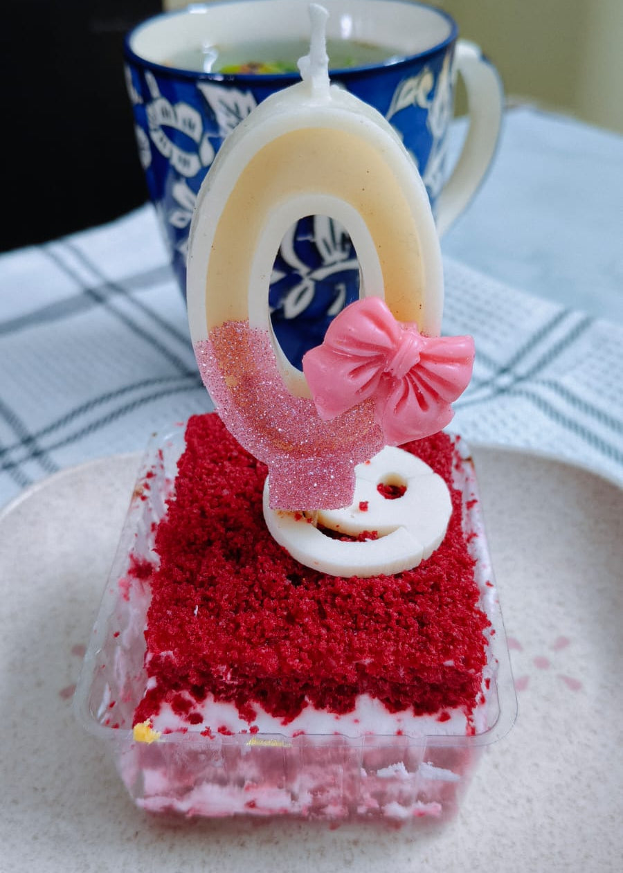 Small red velvet cake with a number 0 candle on a white plate, with a blue mug in the background.